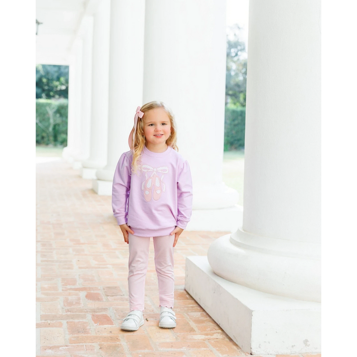 A young girl wearing a That Store lavender French terry pullover with ballet shoe embroidery and matching leggings.