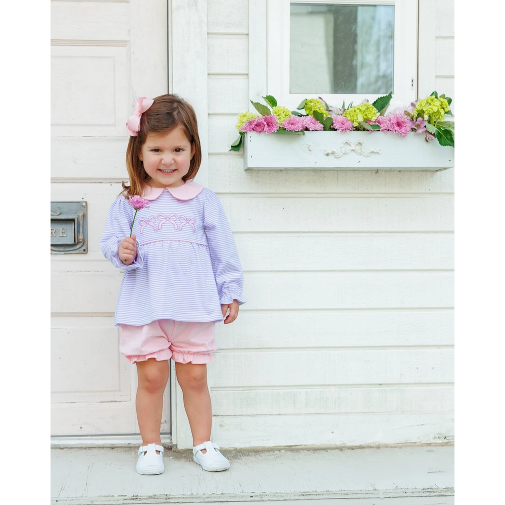A young girl wearing a That Store lavender stripe bloomer set with bow appliqué, standing by a white cottage wall.