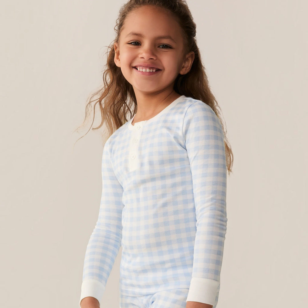A smiling child wearing a That Store light blue gingham Pima cotton long sleeve pajama set with a henley neckline.
