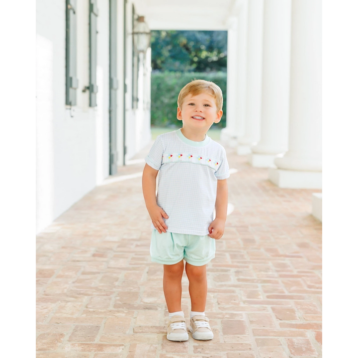 Toddler boy wearing a That Store light blue gingham short set with Mickey balloon ribbon detail on a brick walkway