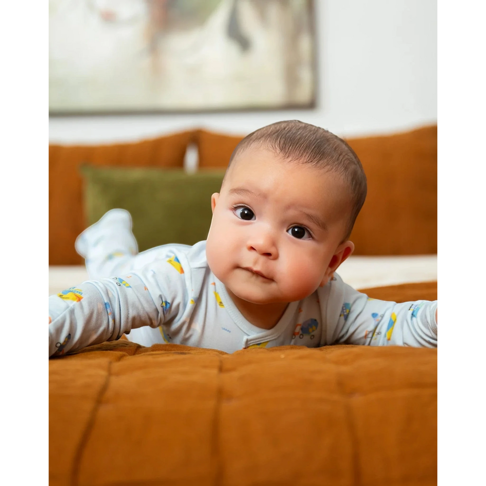 That Store baby wearing a light blue modal magnetic footie with a construction vehicle print while lying on a bed.