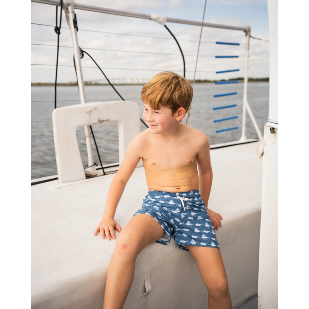 A young boy wearing That Store navy and white fish print Saint Simons swim trunks while sitting on a boat deck.