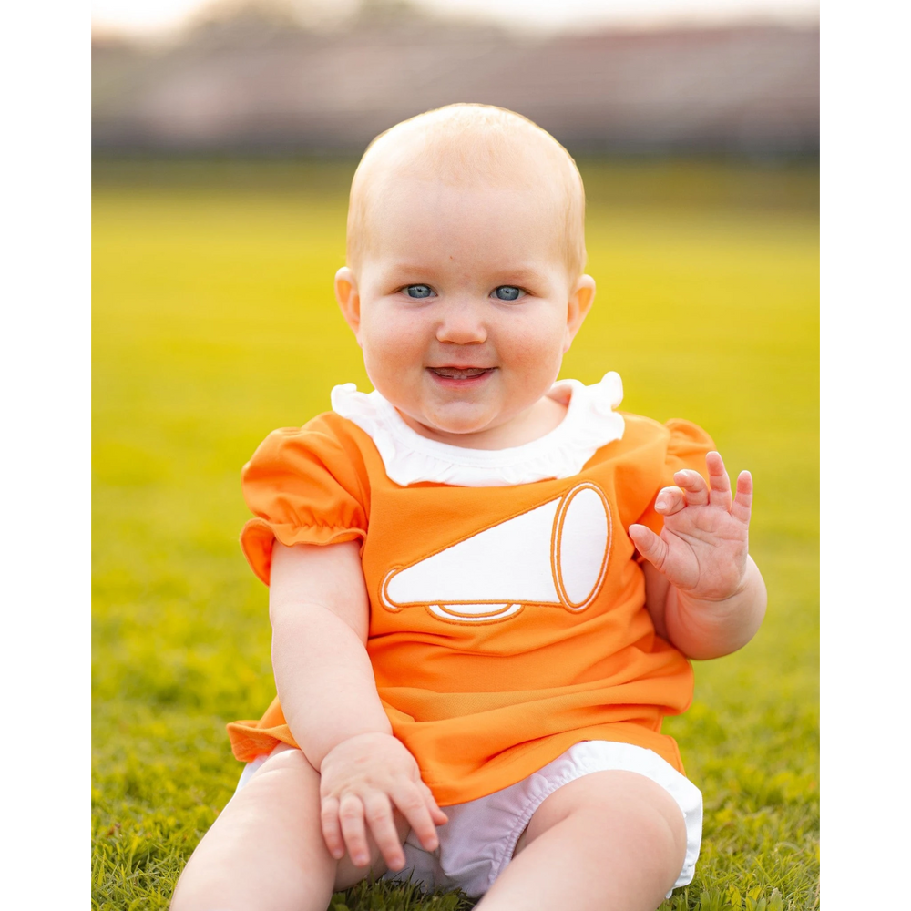 Baby girl wearing an orange That Store bloomer set with a white megaphone applique, sitting on a grassy field.