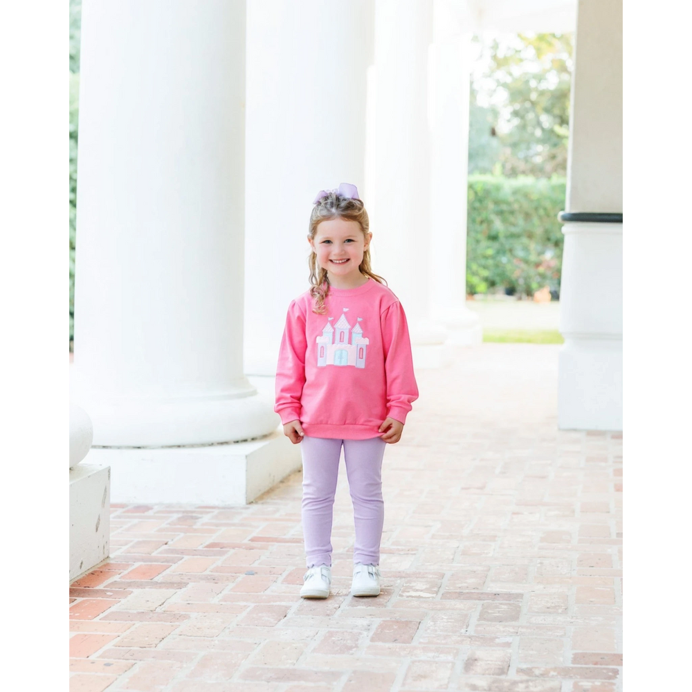 A young girl wearing a That Store pink French terry pullover with a castle graphic, paired with light purple leggings.