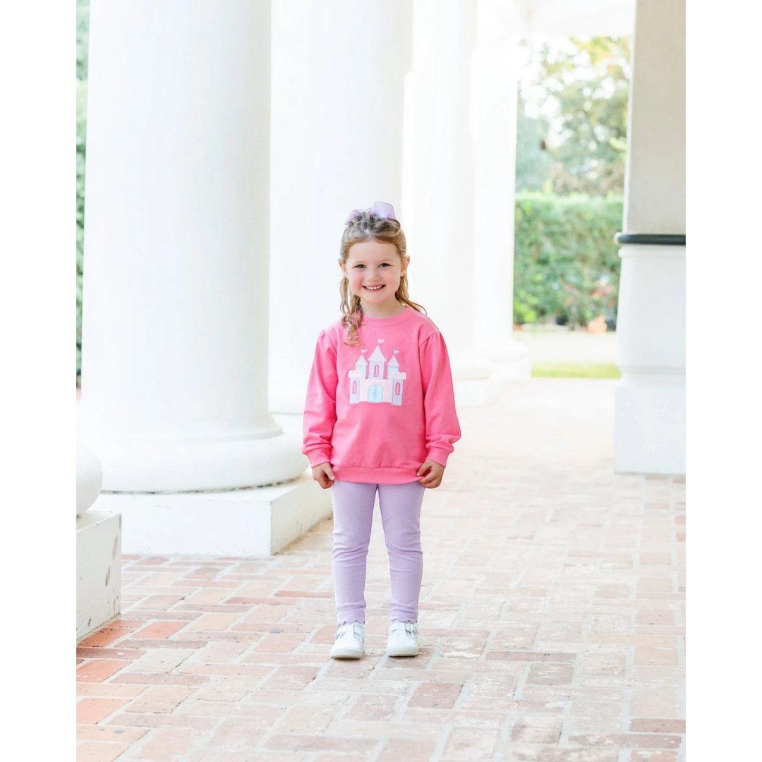 A young girl wearing a That Store pink French terry pullover with a castle graphic, paired with light purple leggings.