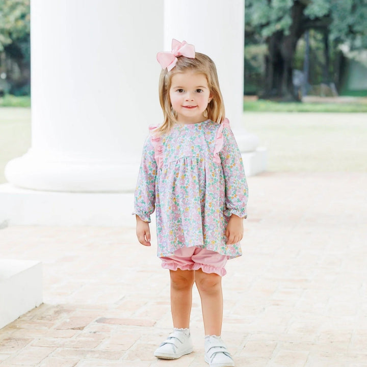 Toddler girl wearing a That Store pink floral bloomer set with ruffled sleeves and a matching hair bow outdoors.