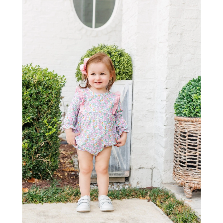 Toddler girl wearing a That Store pink floral bubble romper with ruffled shoulders, standing outdoors.