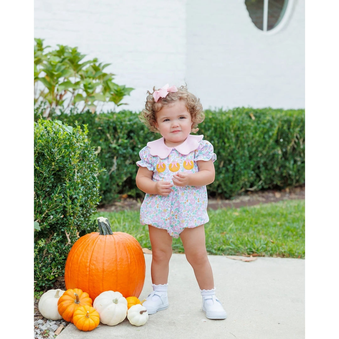 That Store pink floral pumpkin bubble romper worn by a toddler with a pink hair bow standing by pumpkins