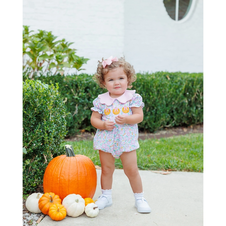 That Store pink floral pumpkin bubble romper worn by a toddler with a pink hair bow standing by pumpkins