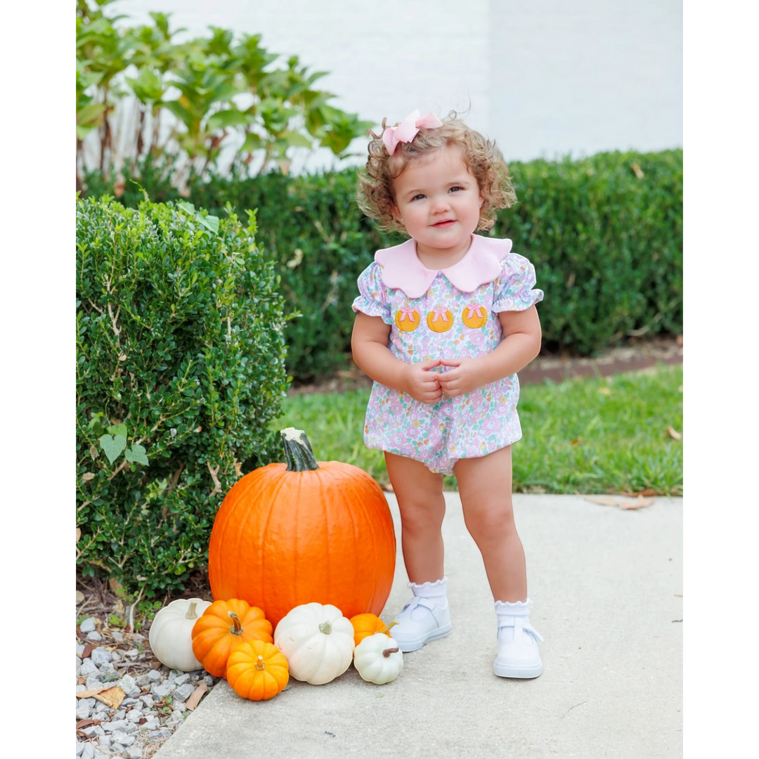 Toddler girl wearing a That Store pink floral pumpkin bubble romper with a Peter Pan collar standing by pumpkins.