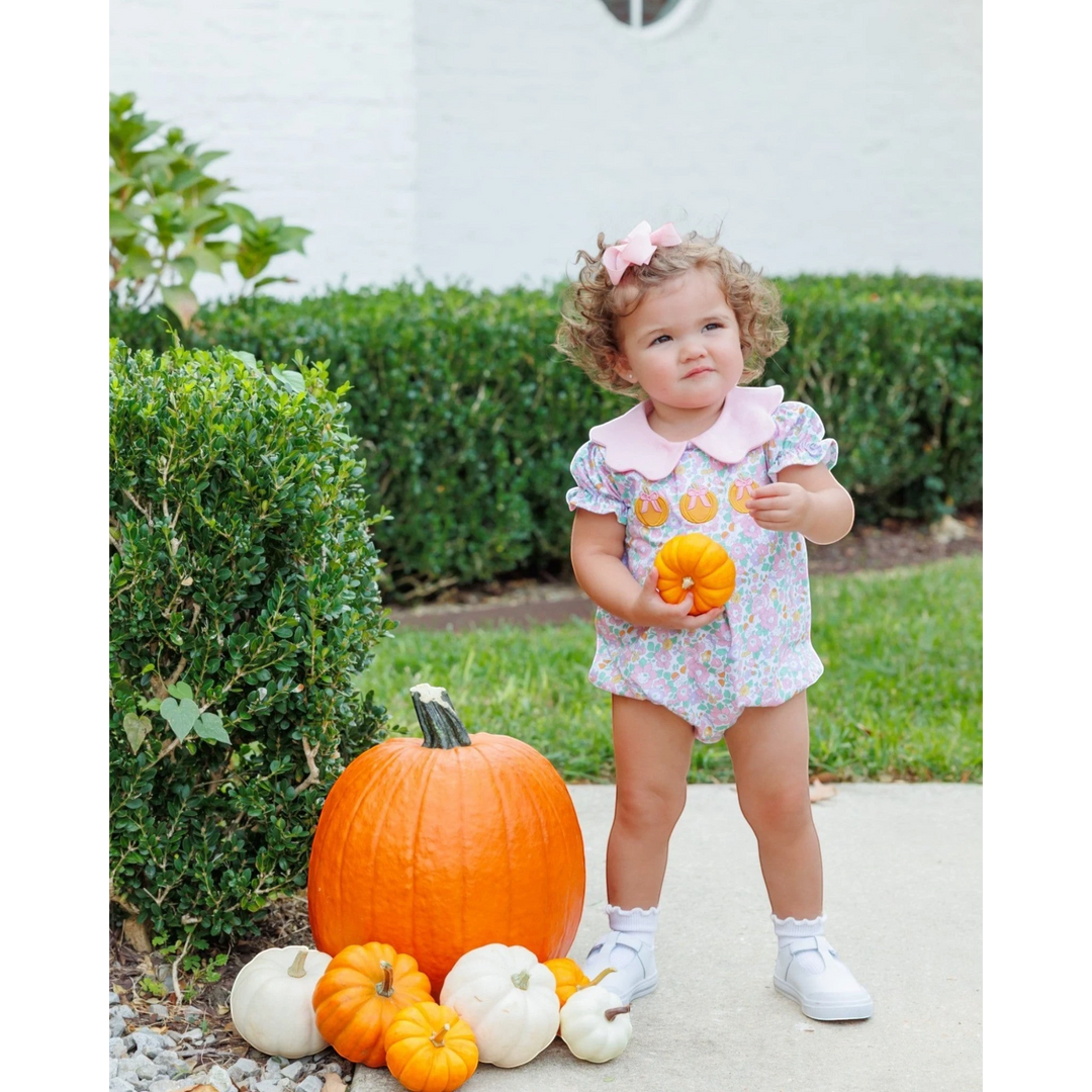 Toddler girl wearing a That Store pink floral pumpkin bubble romper while standing next to pumpkins in a garden