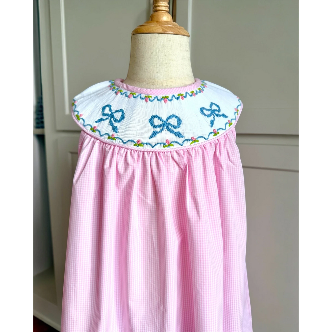 That Store pink gingham sleeveless dress with a white smocked yoke featuring blue embroidered bows and floral detail.