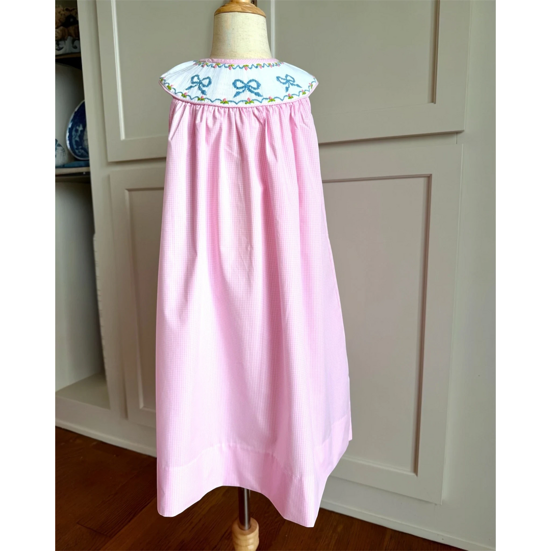 That Store pink gingham sleeveless dress with a white smocked yoke featuring embroidered blue bows and floral details.