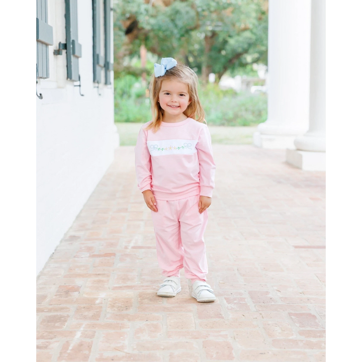 That Store pink girls jogger set featuring delicate floral and bow embroidery, worn by a smiling toddler outdoors.