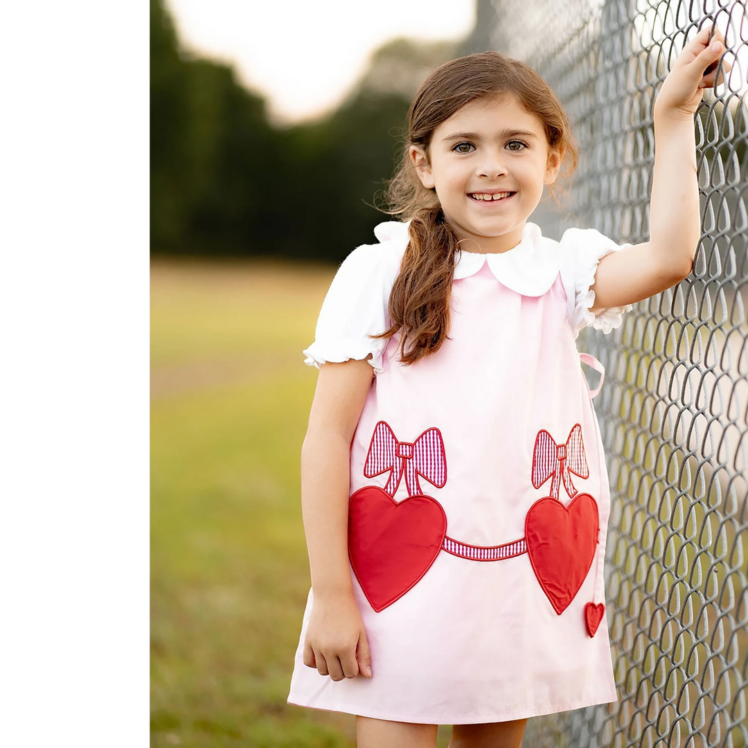 That Store Emma Pink jumper with red heart applique and bow details worn by a young girl standing by a fence