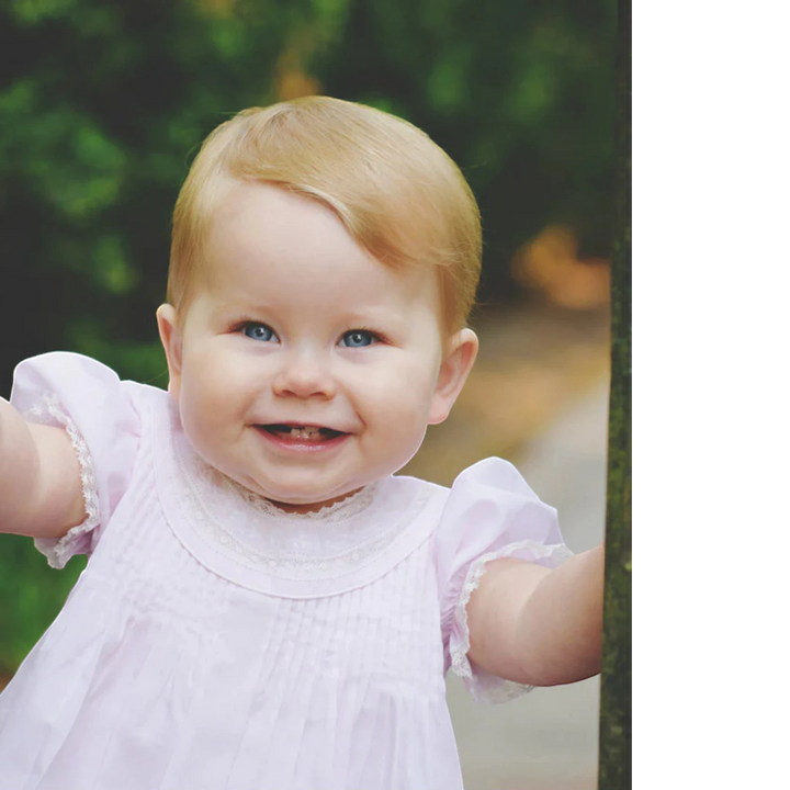 That Store pink embroidered slip dress with lace trim and puffed sleeves worn by a smiling baby girl outdoors