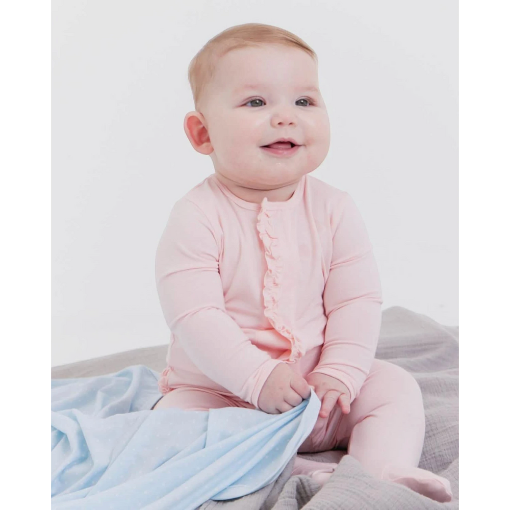 Baby wearing a soft pink That Store modal magnetic footie with ruffle detail, sitting on a grey blanket.