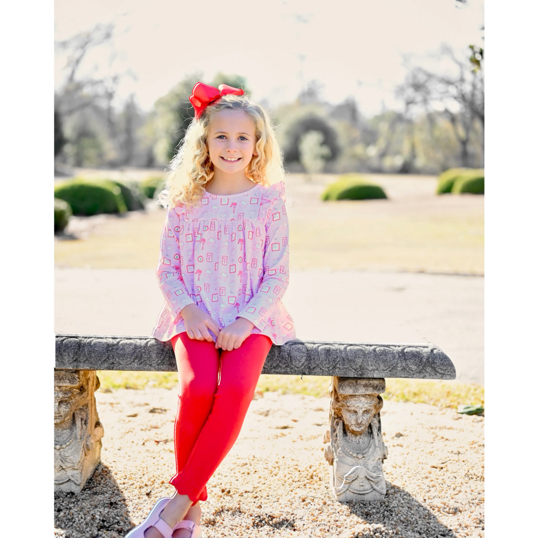 Young girl wearing a That Store pink Letters to Santa print swing top and red knit leggings sitting on a stone bench