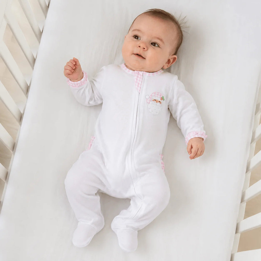 Baby wearing a That Store white Pima cotton footie with pink gingham ruffles and a Santa applique in a crib