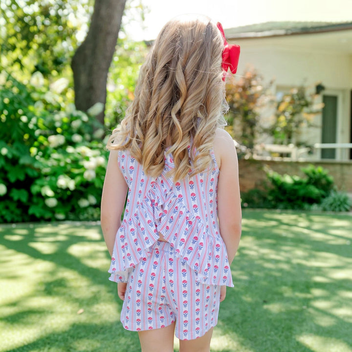 Girl wearing a That Store Poppy liberty floral cotton short set with a red hair bow, viewed from the back outdoors.