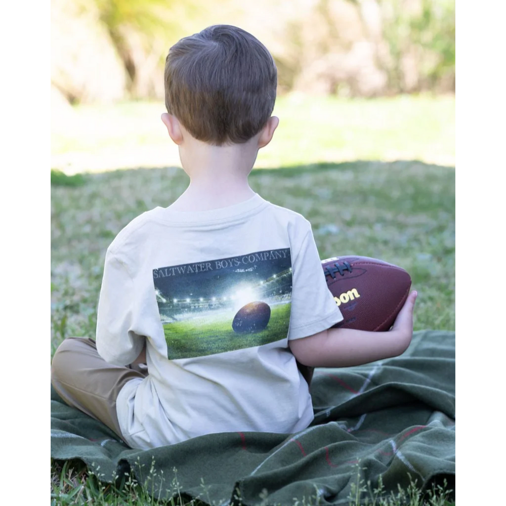 That Store boys' putty graphic tee featuring a football stadium print, worn by a boy sitting on a green blanket.