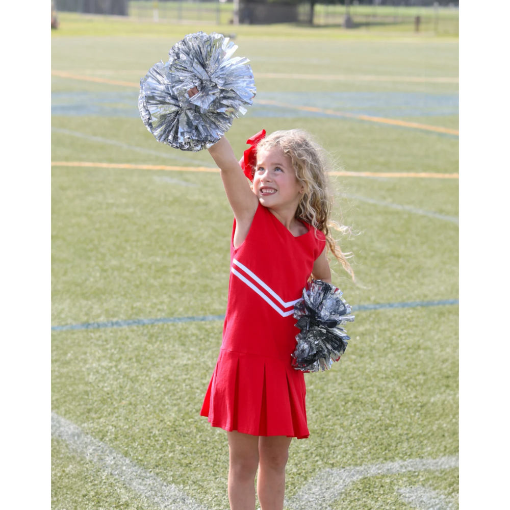 Young girl wearing a That Store red cheer uniform dress with white stripes, holding silver pom-poms on a sports field.