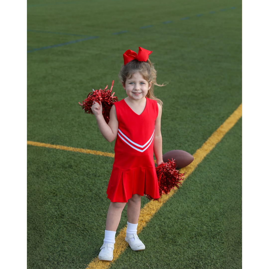 Young girl wearing a That Store red cheer uniform dress with white stripes and holding red pom-poms on a football field