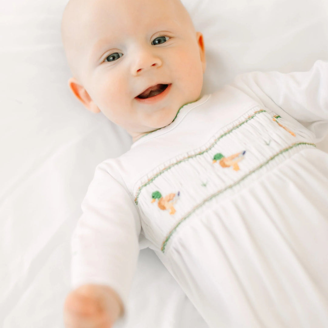 A smiling baby wearing a That Store white Pima cotton footie with a smocked mallard duck pattern on the chest.