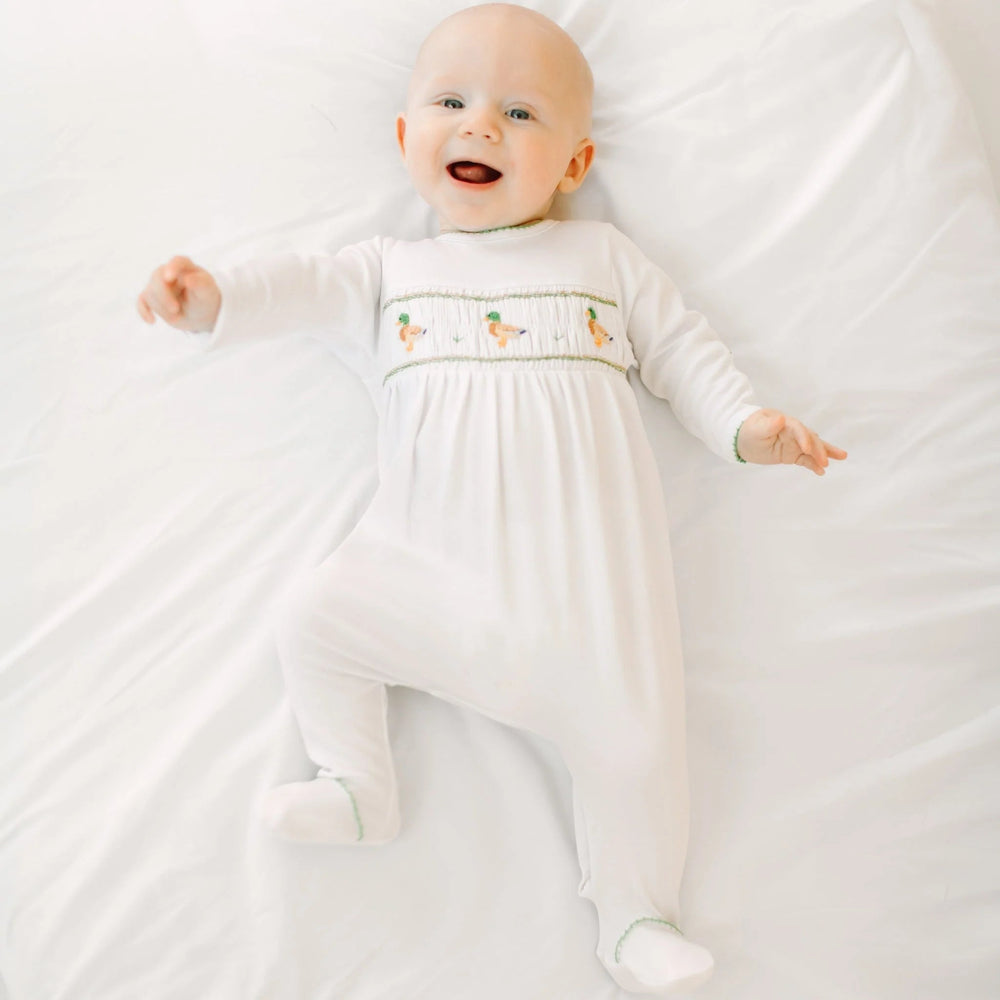 Happy baby wearing a white That Store smocked footie with mallard duck embroidery in soft Peruvian Pima cotton