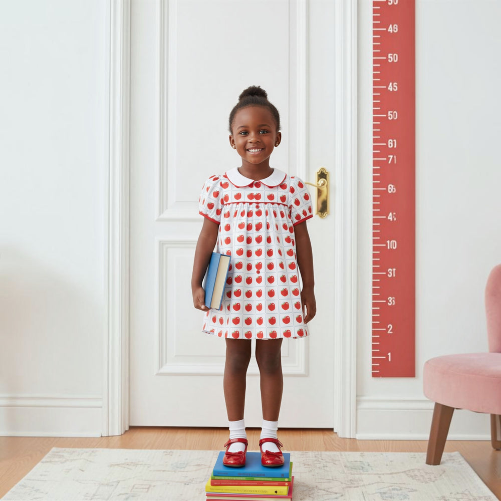 Young girl wearing a That Store Teachers Pet apple print dress with a white collar, standing on a stack of books.