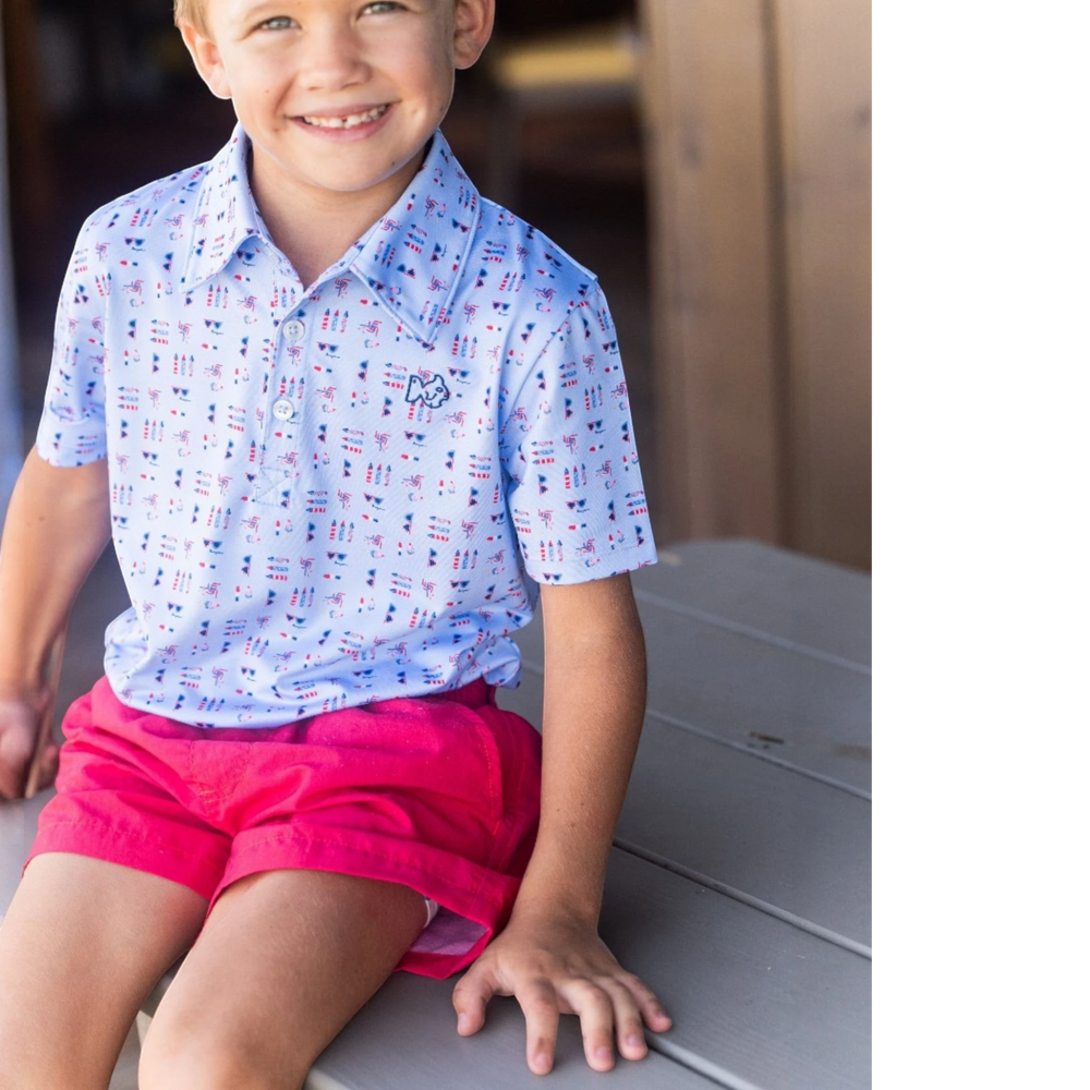 A young boy wearing That Store watermelon performance shorts and a patterned polo shirt while sitting outdoors.