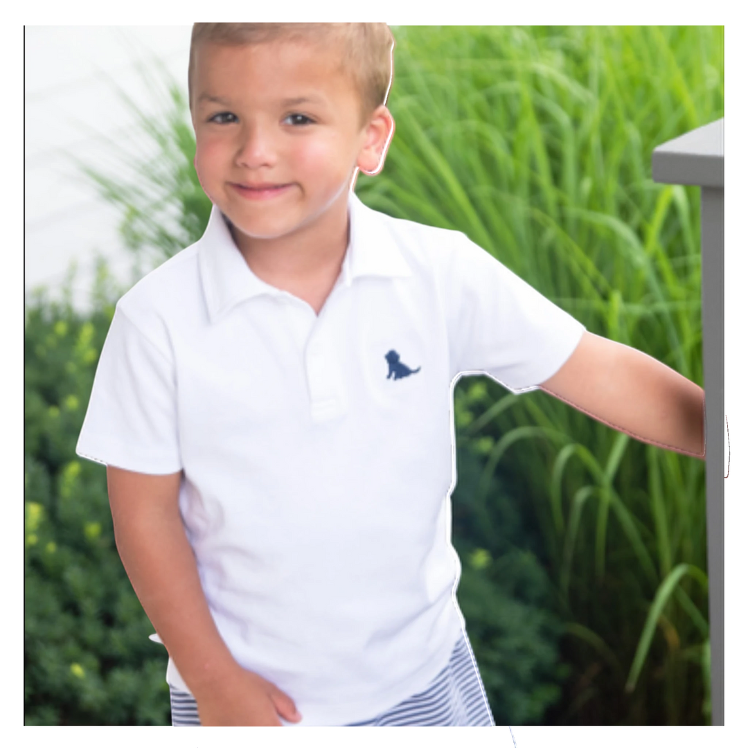 Young boy wearing a That Store white polo shirt with a navy blue embroidered puppy on the chest in a garden setting.