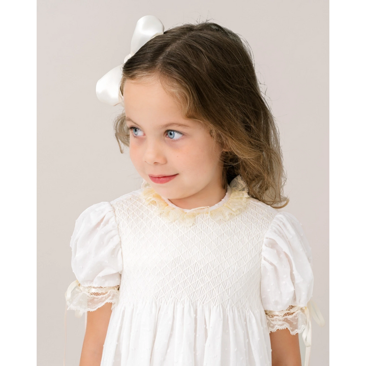 Young girl wearing a That Store white Swiss dot dress with ecru lace trim and a matching white hair bow