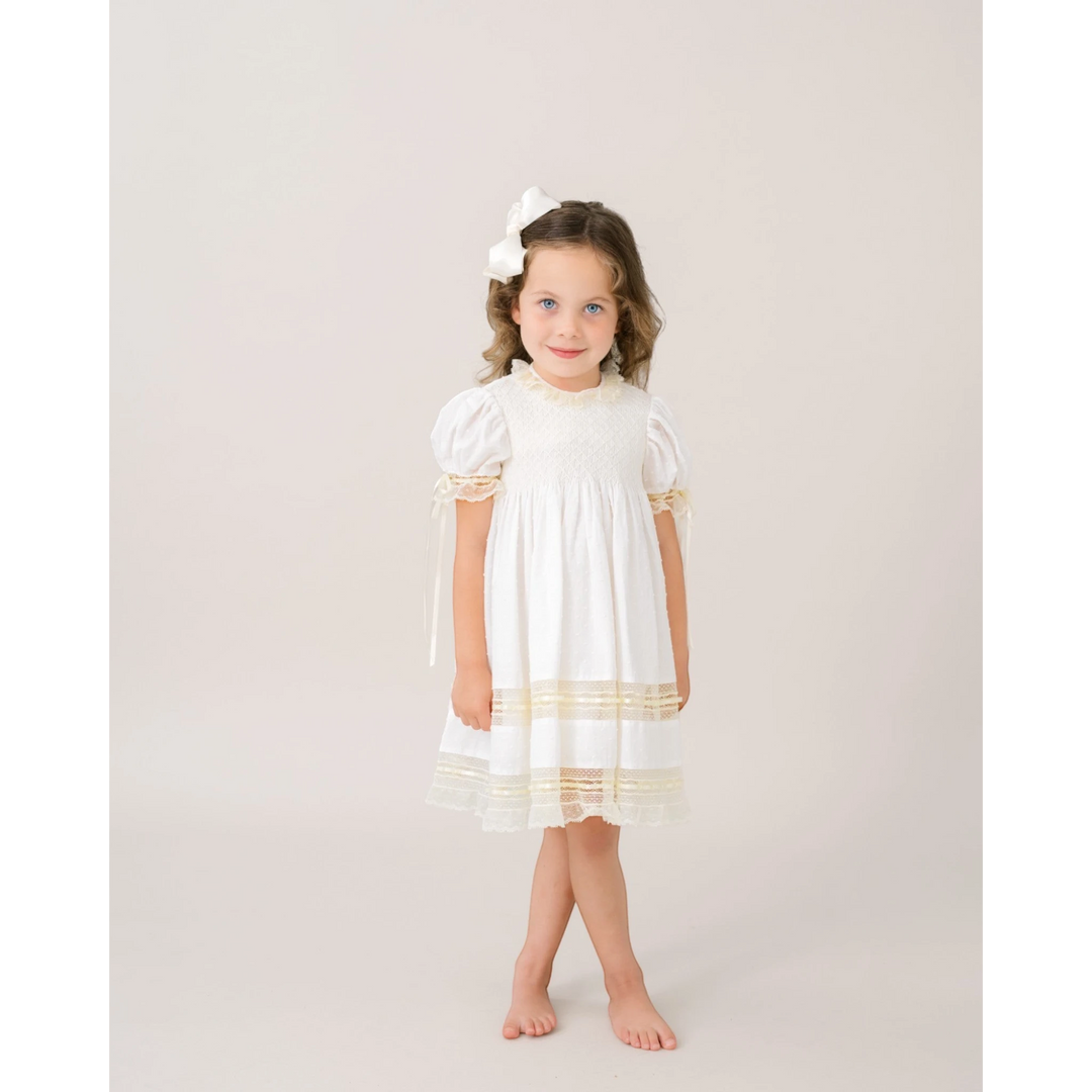 Young girl wearing a That Store white Swiss dot dress with ecru lace trim and a matching white hair bow