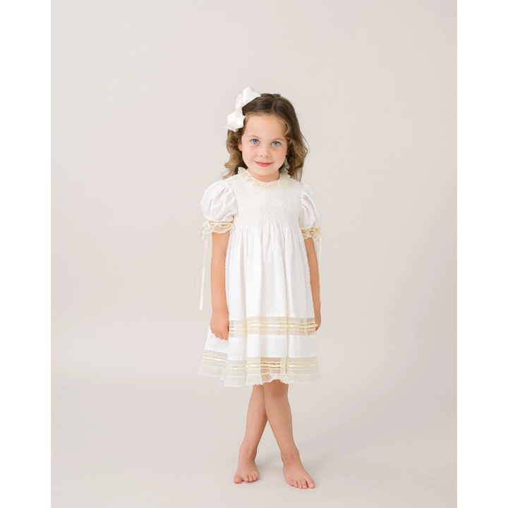 Young girl wearing a That Store white Swiss dot dress with ecru lace trim and a matching white hair bow
