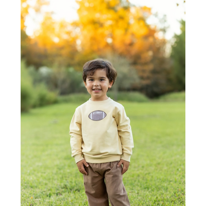 A young boy wearing a That Store yellow French terry pullover with a football graphic, standing in a park.
