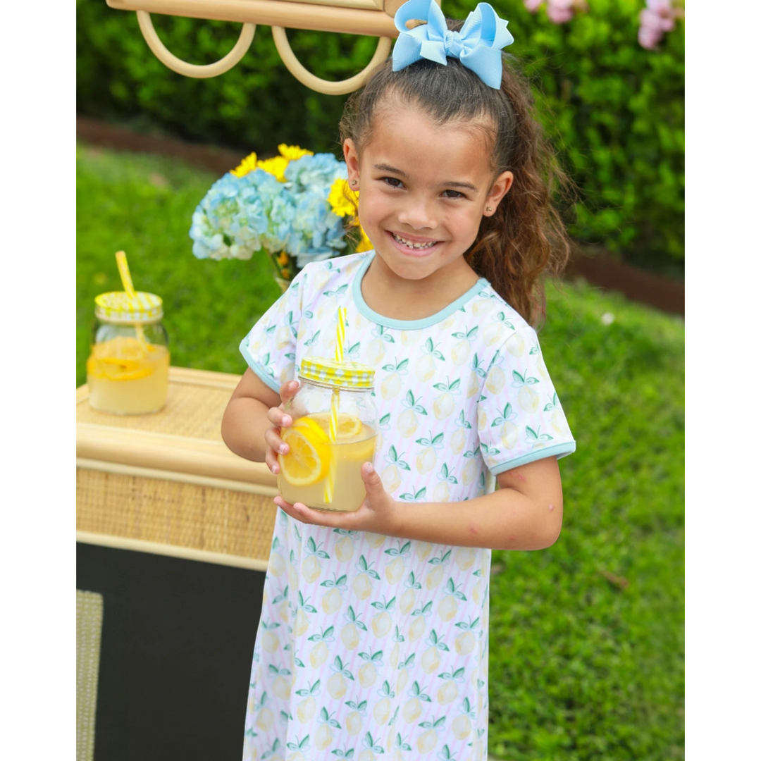 A young girl wearing a That Store yellow lemon-print cotton dress, holding a glass of lemonade in a garden setting.